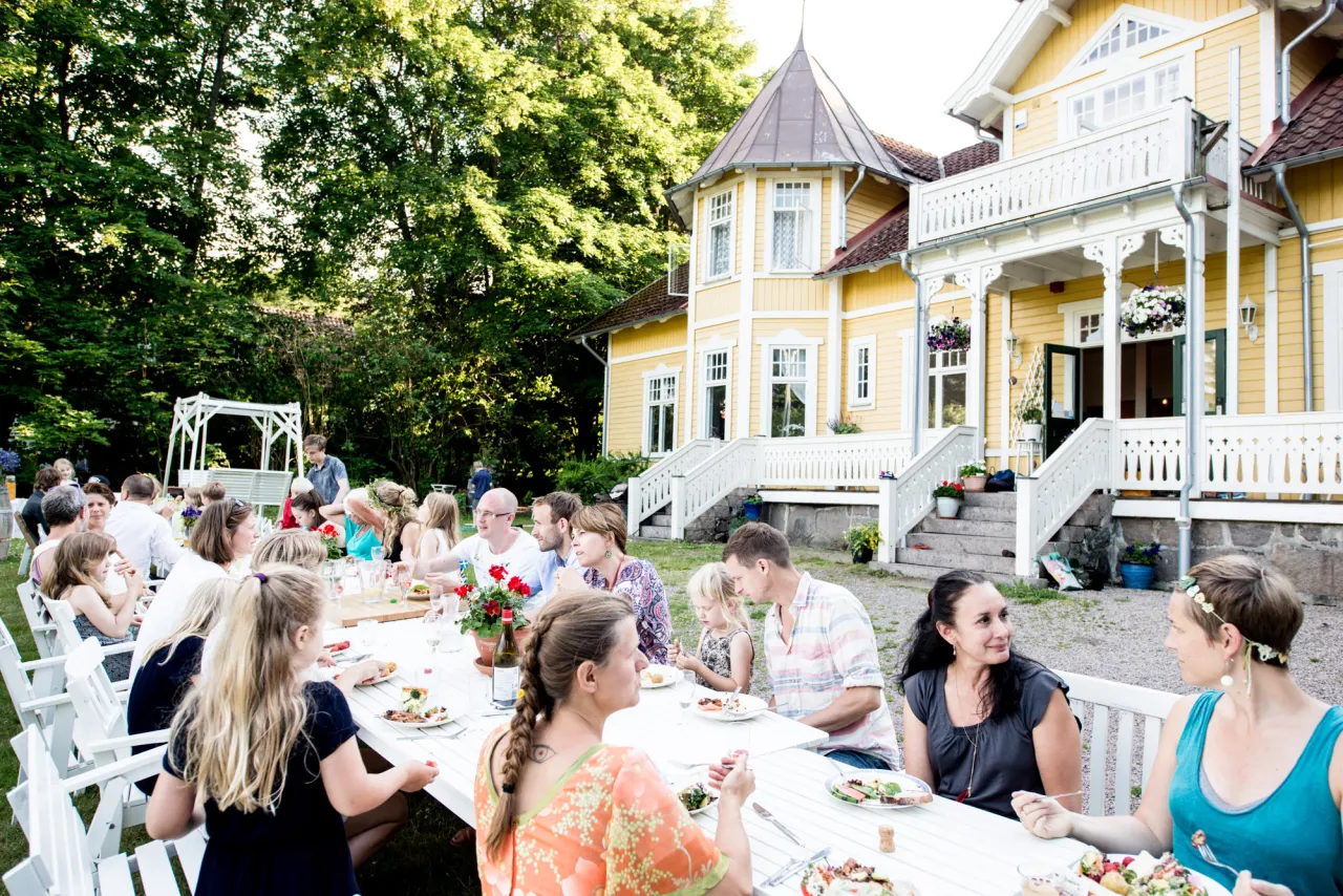 Långt dukat bord med middagsgäster utomhus framför Villa Söderåsen i Röstånga med veranda, omgiven av gröna träd.