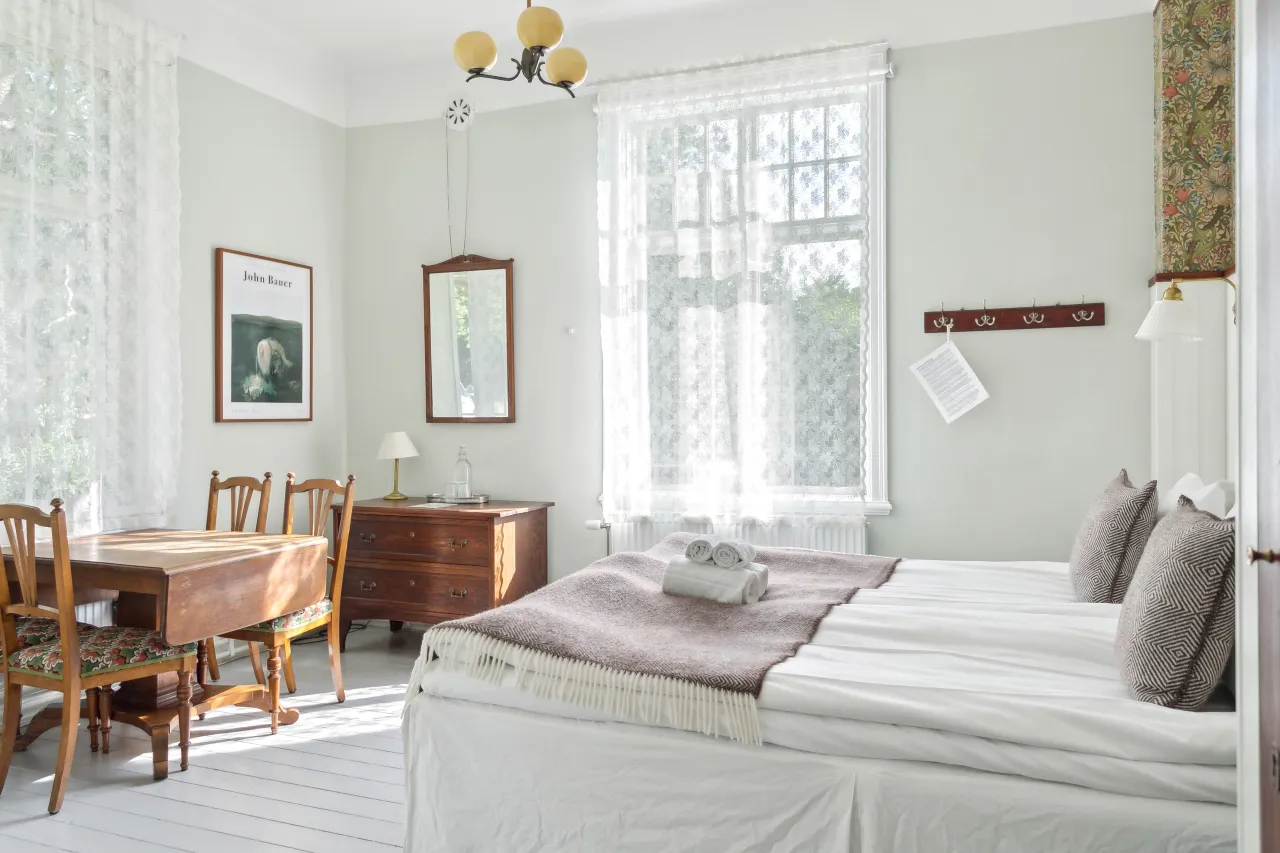 Ljust sovrum med dubbelsäng, vikta handdukar, spetsgardiner vid stora fönster samt bord och byrå i trä.