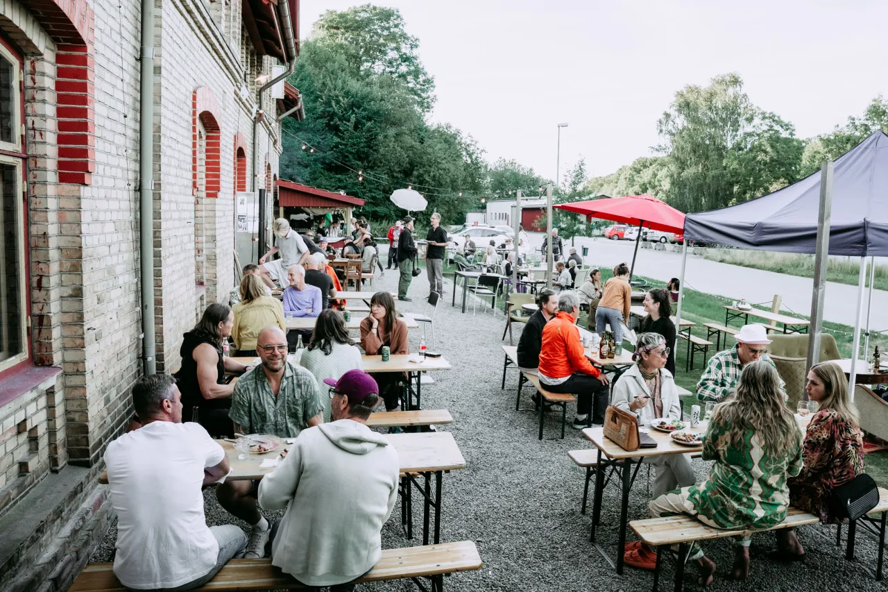 Uteservering med många sittande gäster på baksidan av Stationshuset i Röstånga, bord och bänkar på grus samt parasoller och tält.