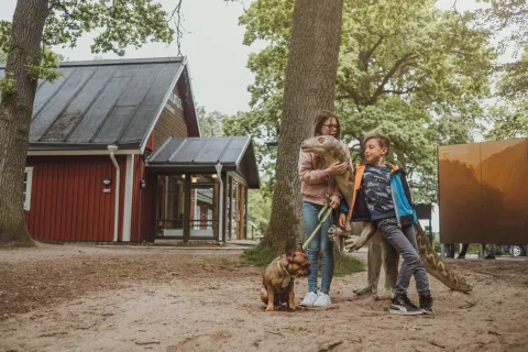 En kvinna och en pojke står vid en dinosauriestaty i en park, med en liten hund framför ett rött hus.