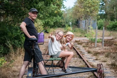 Tre personer på en dressin på en övergiven järnvägsräls i skogsmiljö, två visar tummen upp.