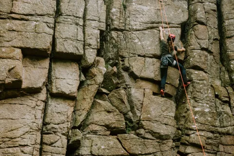 En person med röd hjälm och sele klättrar på en brant klippvägg i Klåveröds strövområde, säkrad med ett orange rep i dagsljus.