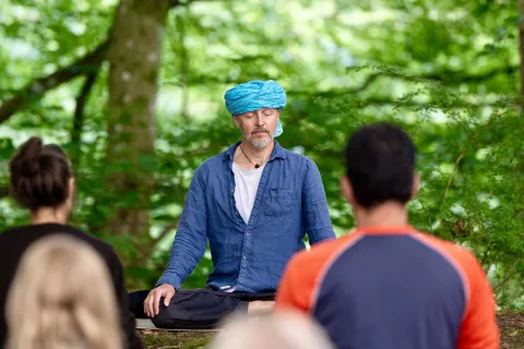 Yogaläraren Sky sitter i korslagd ställning på en matta i en grön skog, med deltagare suddigt i förgrunden.