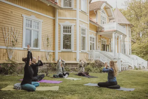 Grupp som gör yoga på mattor på en gräsmatta framför Villa Söderåsen, en gul träbyggnad med vita detaljer och veranda.