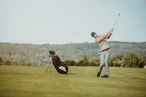 En golfspelare svingar en klubba på golfbanan vid Söderåsen, med en golfbag bredvid och skogsklädda kullar i bakgrunden.
