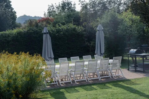 Vit långbord med många stolar och två hopfällda parasoller på stenlagd uteplats i Villa Söderåsens trädgård, med grönskande häck och grill.