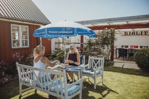 Två personer sitter vid ett bord på en gräsmatta under ett blått parasoll, med hus och uteservering i bakgrunden.