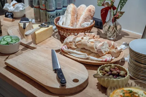 Välkomstfika på Villa Söderåsen. Träbord med skärbrädor, kniv, ost, brödkorgar, gurkskivor, oliver och tallrikar samt en vas med blad.