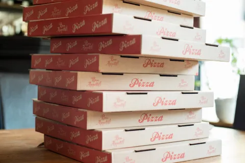 En hög med staplade pizzakartonger i rött och vitt står på ett träbord i ett ljust rum på Roma Pizzeria i Röstånga.