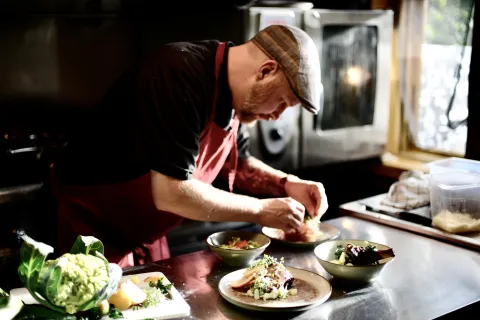 En kock i förkläde arbetar vid en rostfri bänk och lägger upp sallad på tallrikar i ett restaurangkök.