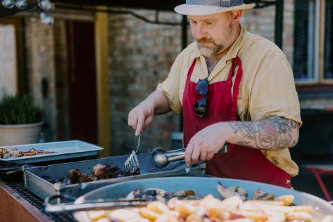 Daniel Green i rött förkläde lagar grillad mat utomhus på Stationshuset i Röstånga med stekspade och tång vid en stor grill.