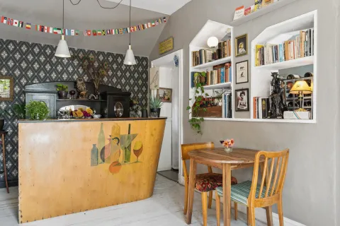 Interiör på stationshuset i Röstånga med träbardisk, mönstrad tapet, hänglampor, inbyggda bokhyllor med böcker, växter och litet bord.