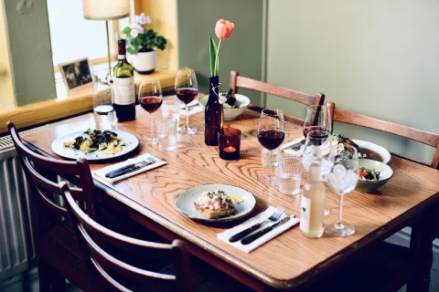 Träbord dukat med tallrikar mat på stationshuset i Röstånga, flera vinglas med rött vin, en vinflaska och en tulpan i vas.